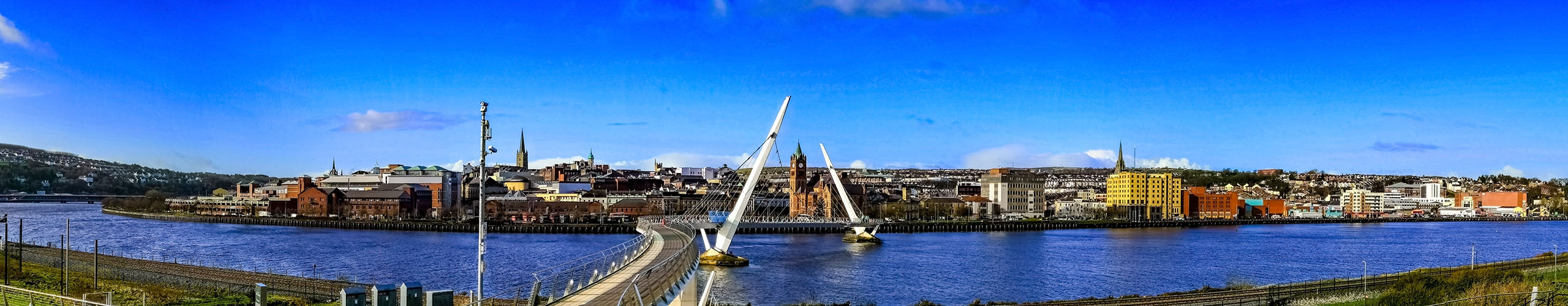 a picture from Ebrington towards the city side showing the river foyle, peace bridge, the city skyline and a blue sky